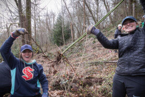Volunteers victoriously holding up a large blackberry plant with root, an invasive plant in the Camp Long forest. Photo by photographer Jamison A. Johnson. 