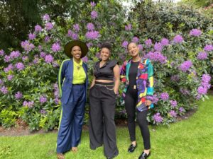 Photo identification description: With a substantially large light-purple colored Rhododendron flowering in the background, three of DNDA’s executive leadership team stand next to each other while smiling gleefully. From left to right, Co-executive director Imani Sims wears a navy-blue suit outfitted with bright highlighter yellow dress shirt and a large-brimmed hat. Board President Shannon Woodard stands with their hands in their pockets in a collared Black silk crop top and wide pants decorated by a gold chain belt. Co-executive director Mesha Florentino tilts their head, posing with one hand on their hip wearing a colorful painterly blazer and Black pants and shirt.