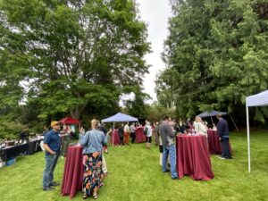 Photo identification description: With towering evergreen trees on an overcast day and a perfectly manicured lawn, many guests converse with each other while standing at multiple circular bar tables outfitted with maroon-colored tablecloths. A few tent coverings are scattered throughout, and to the left is a vendor table from Footprint Wine Tap and Adrice Wines. 