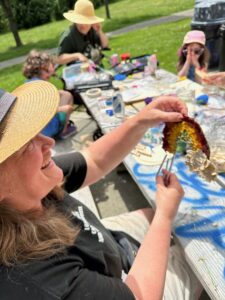 Person proudly shares her mixed media rainbow wind chime- made with all natural materials. In the background we have joyful children and the teaching artist letting their creativity flow in the sunshine.