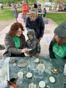 A young student and his grandmother curiously peek at his plaster sculpture that has been drying while he was playing. The teaching artist excitedly helps with the big reveal in a beautiful intergenerational moment.