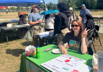 A picture of our End of Program Celebration is featured. We see a group of people surrounding different tables and a blue canopy outdoors at Roxhill Park, speaking to each other in the background. A person at the green table with a DNDA t-shirt smiles and holds up a peace sign.