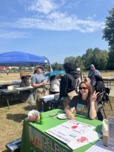 A picture of our End of Program Celebration is featured. We see a group of people surrounding different tables and a blue canopy outdoors at Roxhill Park, speaking to each other in the background. A person at the green table with a DNDA t-shirt smiles and holds up a peace sign.  