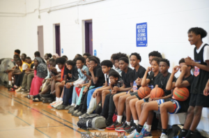 A group of excited young athletes sit together on the sidelines of a gym, eagerly watching a basketball event and preparing to join the action.