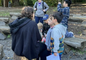 Each has a scavenger hunt made up of a small group of younger and older people surrounding Environmental Education Guides. The Guides are prompting the groups to learn about the site and work to figure out a way for the group to solve their location clues. The group looks and listens to the Guide giving them more information about owls and centipedes to continue on their mission around the Park.