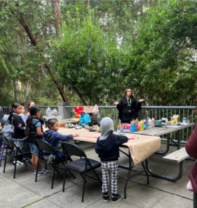 A photo of Nicole Parish-Andrews from Seattle Parks and Recreation’s Environmental Education Team. Nicole, dressed in a black cape, speaks to a group of young people surrounding tables at Camp Long.