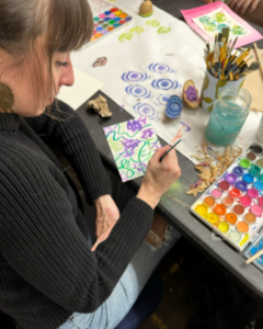 Woman painting purple flowers with water colors. In the background is a rainbow art pallet next to art brushes and other art work. 