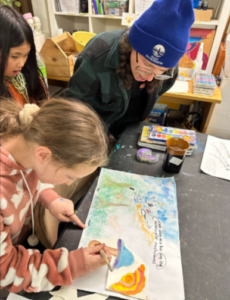 A group of people collaborating on a colorful art project, with one of the teaching artists guiding a participant painting a mushroom design on a vibrant, nature-themed piece.