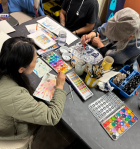 Three people creating art at a table with watercolors, brushes, and sketchbooks in a collaborative, creative setting.