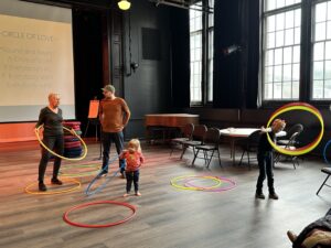 Two adults are speaking to each other as two young kids play with colorful hula hoops, trying to twirl them.