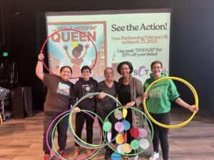 Five people are in front of a projector screen holding colorful hula-hoops in the air together. One person, Gloria, is holding our community art project about the symbolism of a circle.