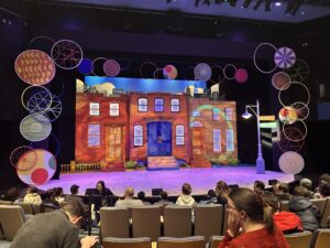 A photo of the Seattle Children's Theater stage set up for the opening night performance The Hula Hoopin' Queen. It is surrounded by colorful hula hoops, bright lights hit the stage where there a mock Harlem, New York neighborhood. Audience members with their children look on in excitement.