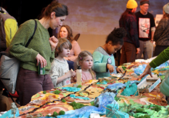 A group of event attendees stand around a 3D map of Longfellow Creek made of recycled materials. One participant is adding to it using glue and small objects, and the others are looking at the map and at other participants standing around the table.