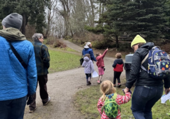 A photo of a group of people walking along the trails of Camp Long. Nicole Parish-Andrews from Seattle Parks and Recreation’s Environmental Education Team leads the group. Nicole, dressed in a blue coat and white beanie hat looks towards where a young participant in the group is pointing.