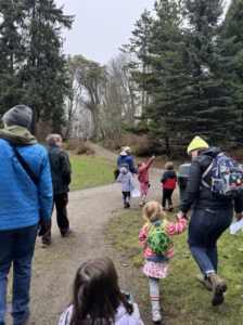 A photo of a group of people walking along the trails of Camp Long. Nicole Parish-Andrews from Seattle Parks and Recreation’s Environmental Education Team leads the group. Nicole, dressed in a blue coat and white beanie hat looks towards where a young participant in the group is pointing. 