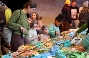 A group of event attendees stand around a 3D map of Longfellow Creek made of recycled materials. One participant is adding to it using glue and small objects, and the others are looking at the map and at other participants standing around the table. 