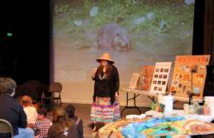 Suquamish Nation elder and artist Barbara Lawrence stands in front of a group of people sitting on the floor and in folding chairs, smiling while telling a story. She is wearing a large woven hat, black top, and colorful skirt. Behind her, there is a large projection of a beaver in a creek. 