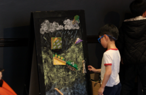 A child at SPU’s Raindrop Run game places a wooden model of the Space Needle on a small chalkboard. The chalkboard also has a small wooden mountain, a hillside with grass, and a sloped roof. 