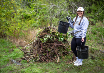 Girl in white hate wearing a grey hoodie and black gloves carrying two black buckets next to a pile of cut down branches.