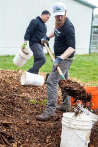 Two older volunteers wearing Nucor gear helping lay down mulch from a white bucket with shovels.