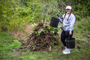 Girl in white hate wearing a grey hoodie and black gloves carrying two black buckets next to a pile of cut down branches.