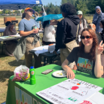 A picture of our End of Program Celebration is featured. We see a group of people surrounding different tables and a blue canopy outdoors at Roxhill Park, speaking to each other in the background. A person at the green table with a DNDA t-shirt smiles and holds up a peace sign.