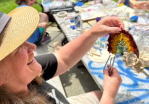 Person proudly shares her mixed media rainbow wind chime- made with all natural materials. In the background we have joyful children and the teaching artist letting their creativity flow in the sunshine.