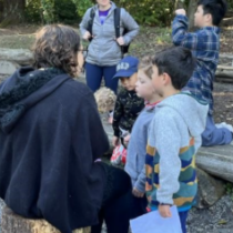 Each has a scavenger hunt made up of a small group of younger and older people surrounding Environmental Education Guides. The Guides are prompting the groups to learn about the site and work to figure out a way for the group to solve their location clues. The group looks and listens to the Guide giving them more information about owls and centipedes to continue on their mission around the Park.