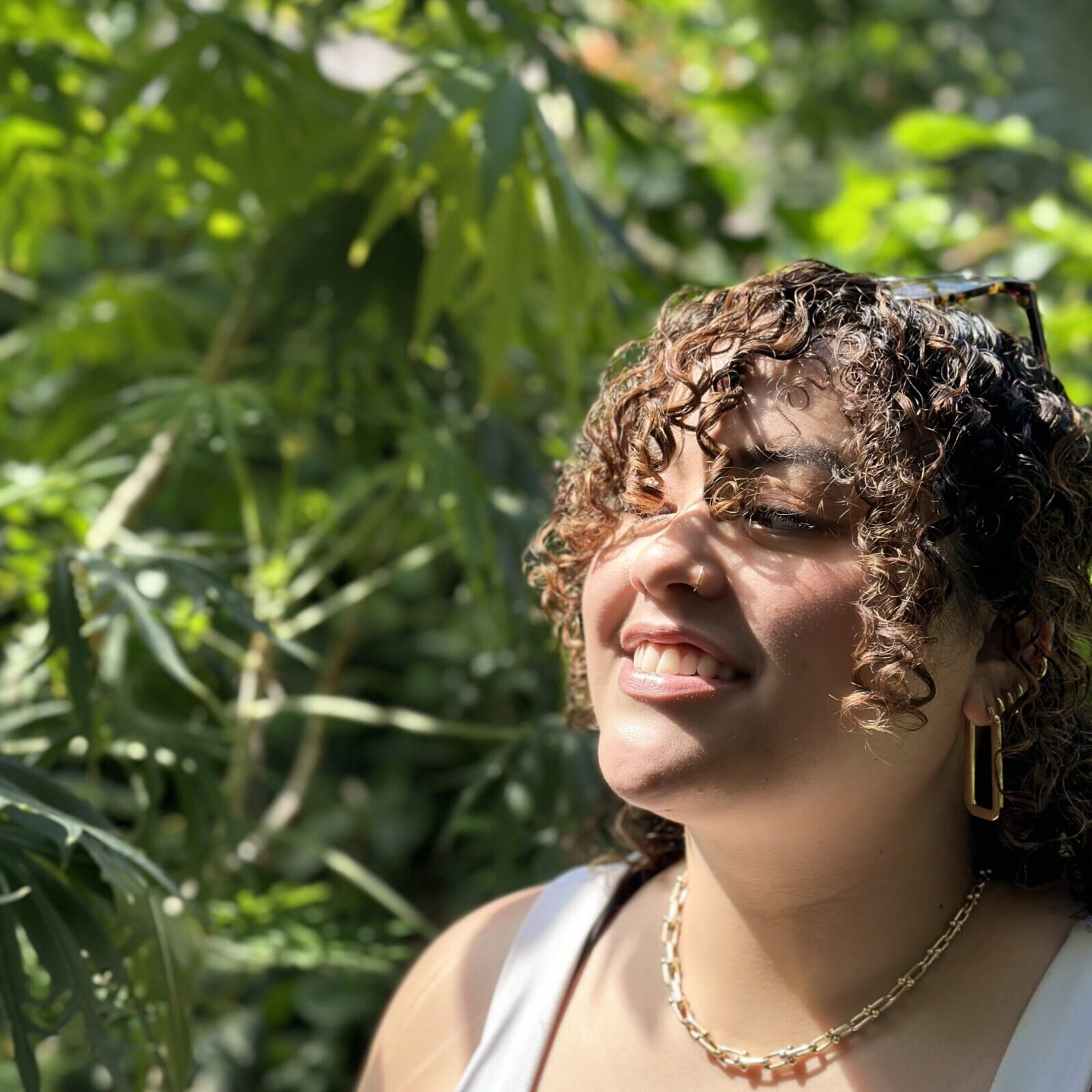 Ranny, a light-skinned Southeastern Asian and Black woman with curly brown bangs is facing outward and smiling in the sunshine with a background of big green leaves. They're wearing a white tank top and gold chain necklace.